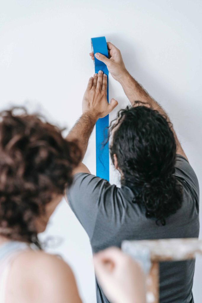 Woman behind the a man wearing gray shirt sticking blue painter's tape on the white wall