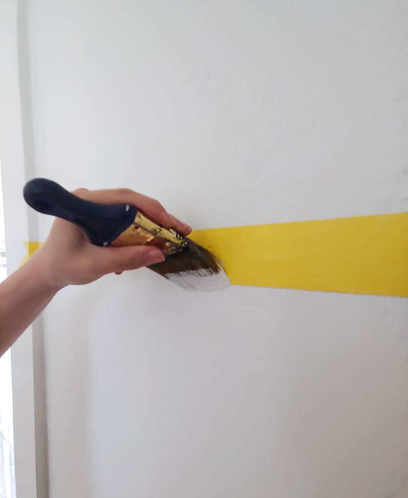 Hand of a woman holding a paintbrush covering the yellow painter's tape horizontally taped on the white wall to seal the tape edge and prevent bleeding or seeping under the tape