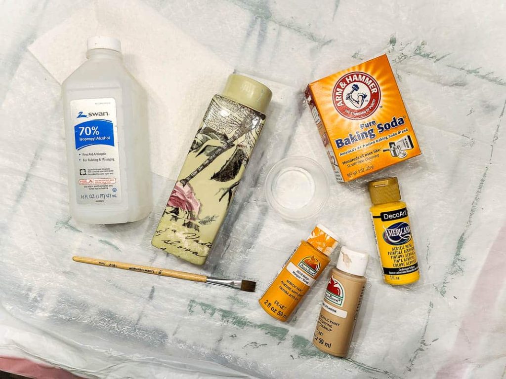 Supplies for painting vase laid on table with bottle of rubbing alcohol, floral vase, paint brush, cup, box of baking soda and three bottles of craft paint