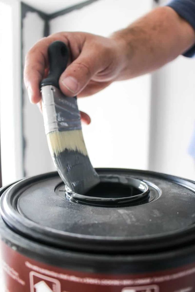 Man holding an angeled paint brush with a short handle pulling out of the paint bucket with gray paint