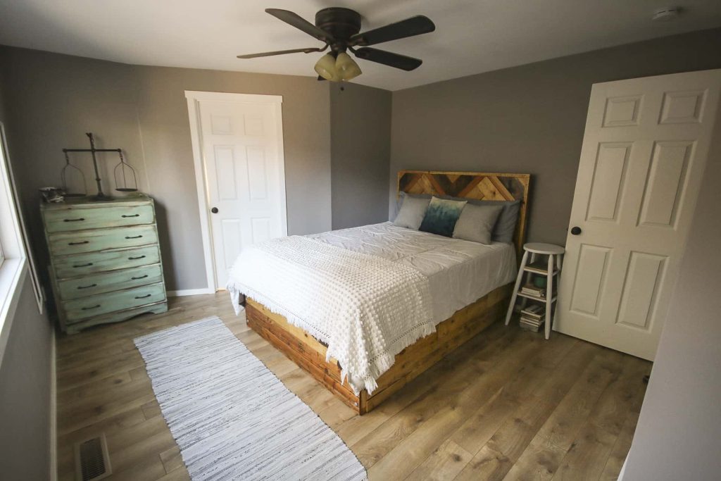Master bedroom side view of the bed, rug, wooden side table and a farmhouse drawer with decor on top beside the bathroom white door and a ceiling fan with chandelier and a painted wall with fashion Gray by Behr 