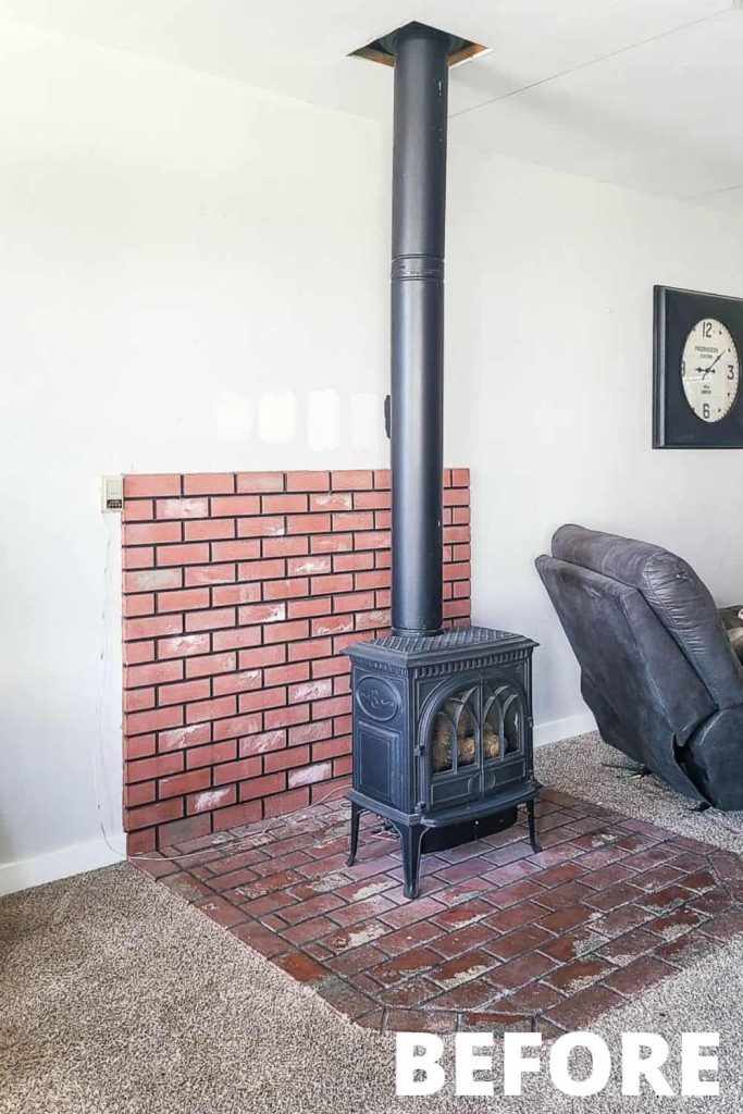 A black freestanding gas fireplace sits in the center of a brick-paved hearth area, with a large chimney that extends into the ceiling. There's a black leather recliner sitting to the right of the fireplace.