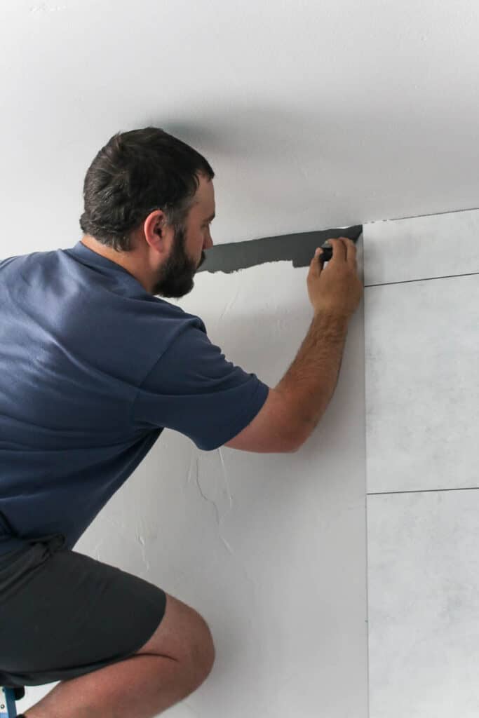 Man standing on top of a ladder wearing blue shirt holding a trim paint brush and painting a straight line with dark gray paint where the wall meets the ceiling