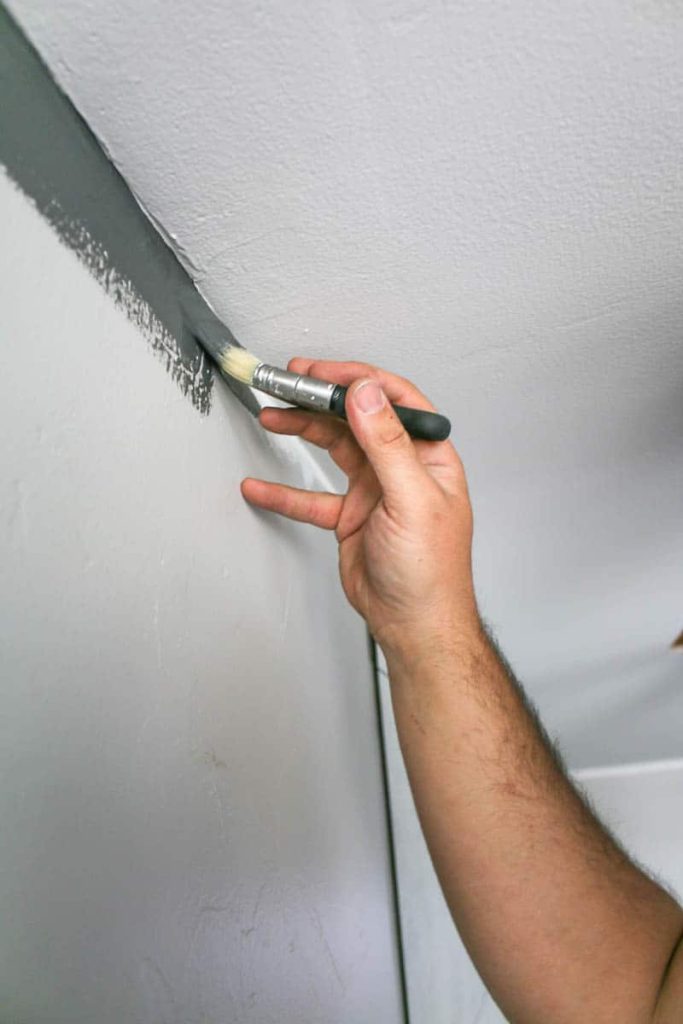 Man holding an angeled paint brush painting the ceiling edge with a straight line with gray paint without using painter's tape