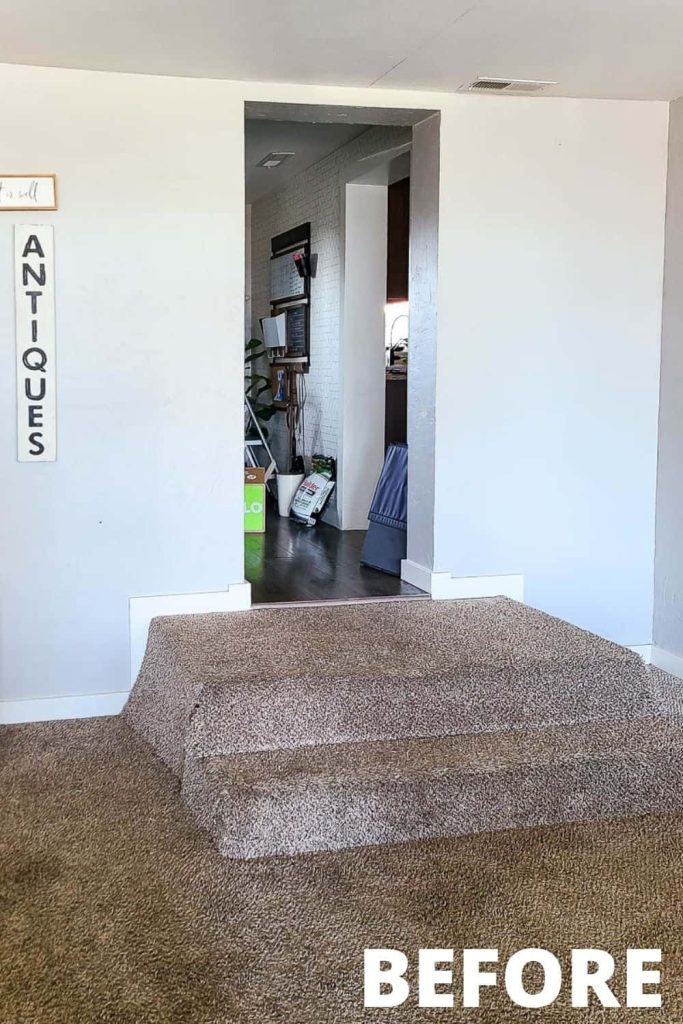 An image of an entryway into a living room with two carpeted steps leading down into the room. The walls have been painted a light color, and there's a vertical decorative wood sign hanging on the wall that says "antiques"