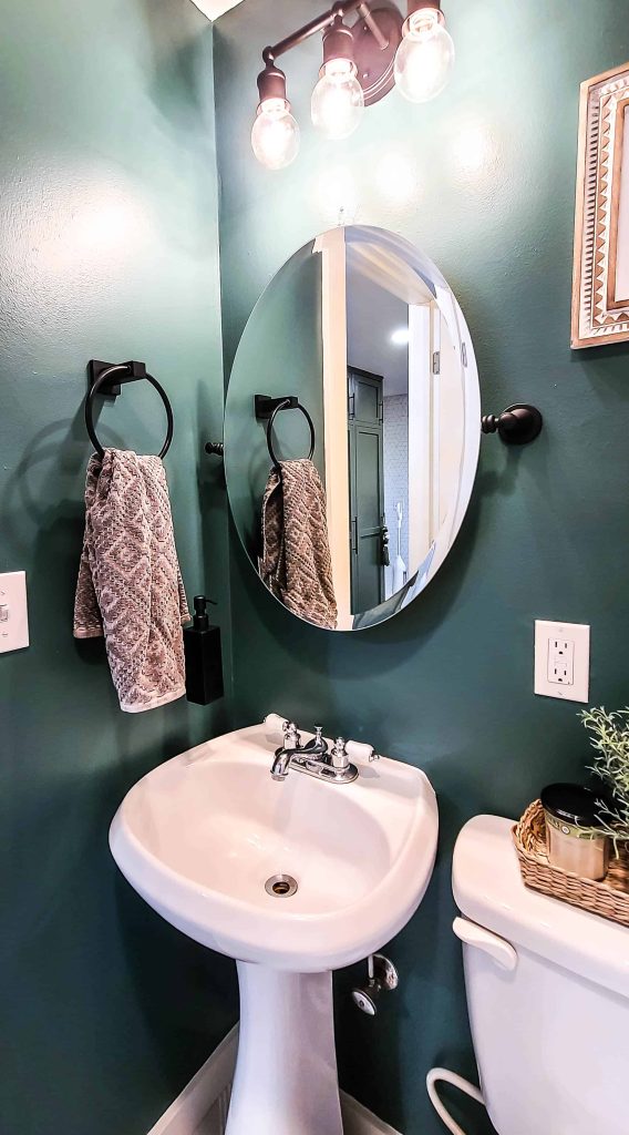Newly painted matte black vanity mirror, dark green painted walls, towel holder, black wall mounted soap bottle, small bathroom sink and light fixtures after the spray paint mirror update