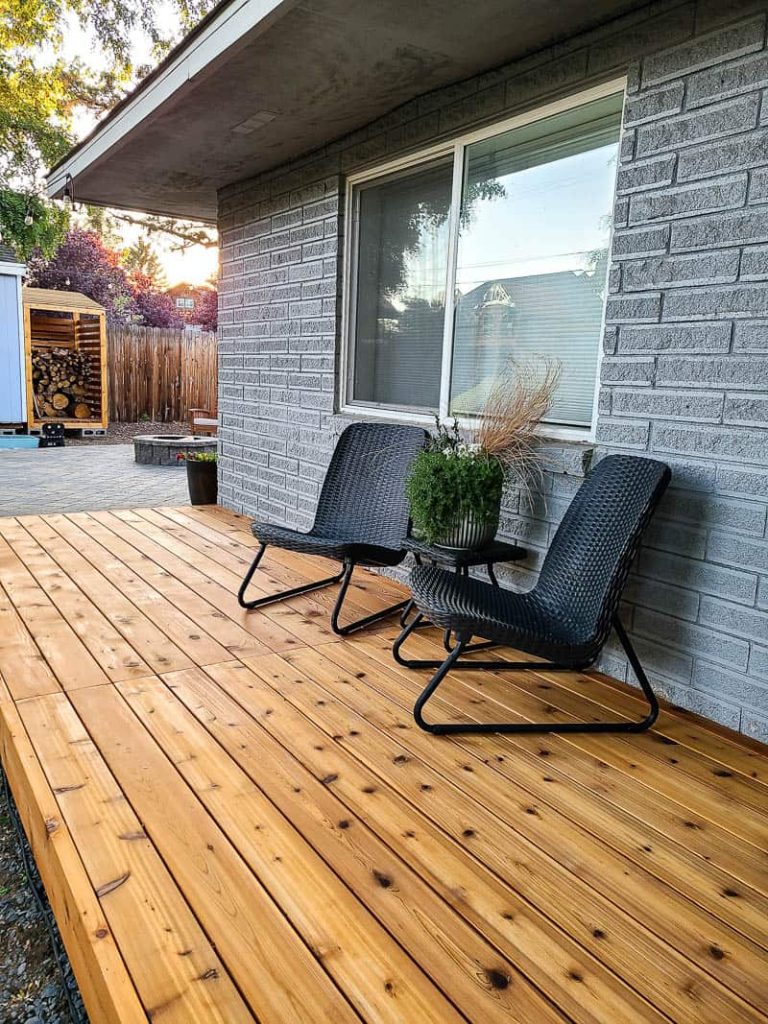 Side view of the backyard with wood deck and black chairs, center table and plants as decor