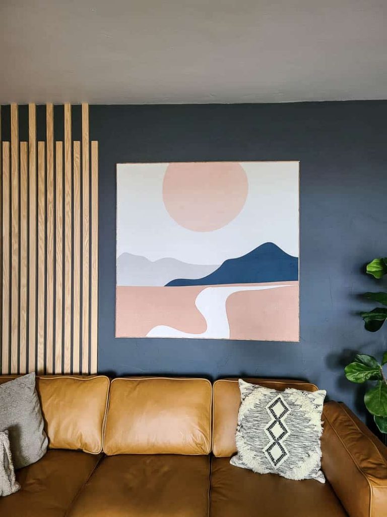 Fabric wall art on the living room hanging on the wall beside wood slat wall above the tan leather couch and boho throw pillows