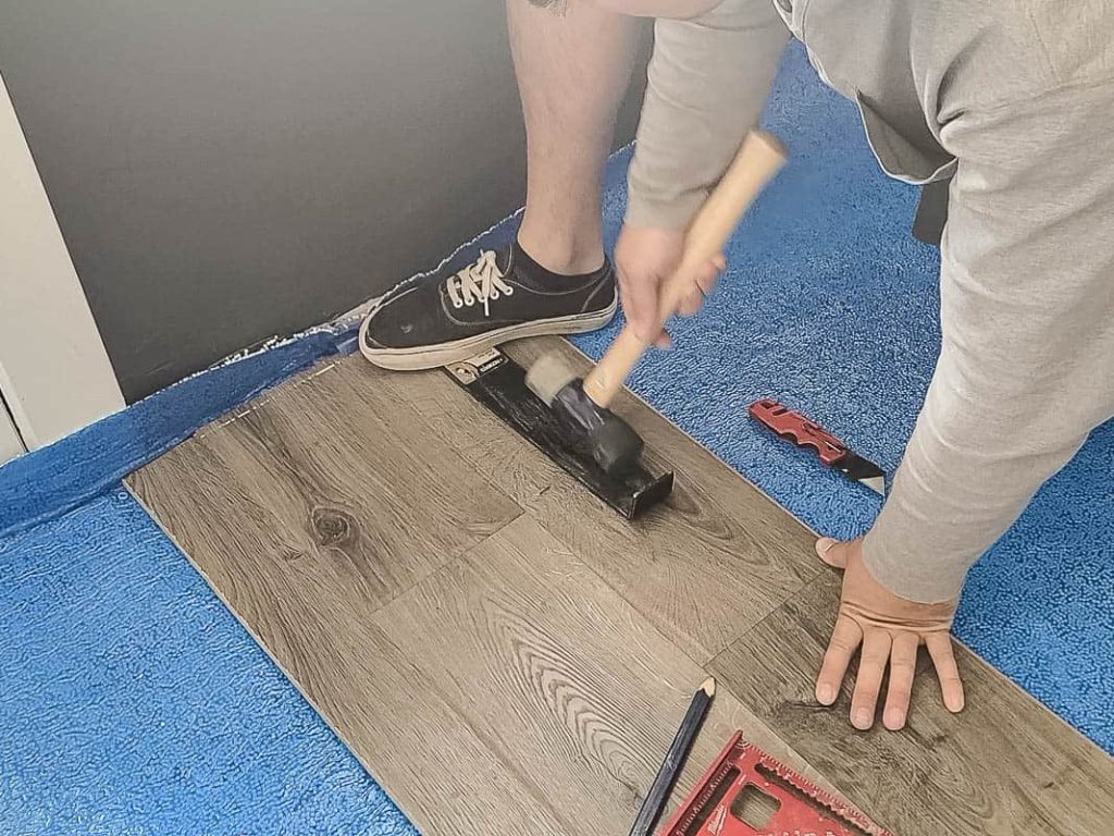 Man attaching the laminate flooring on the edge of the room with hammer and pull bar with other materials