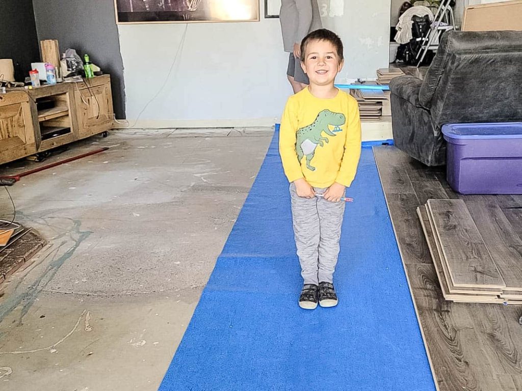 Boy wearing yellow sweater and pants standing on the standing in the underlayment and undone laminate flooring and uncovered concrete floor