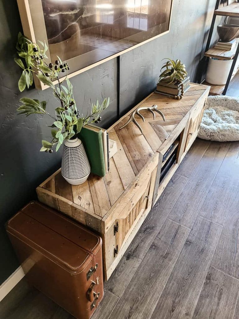 Above shot of TV console made with pallet wood with herringbone top with vintage suitcase standing on its side next to it and a vase used a bookend with greenery, a deer antler and stack of books with an air plant and beads.