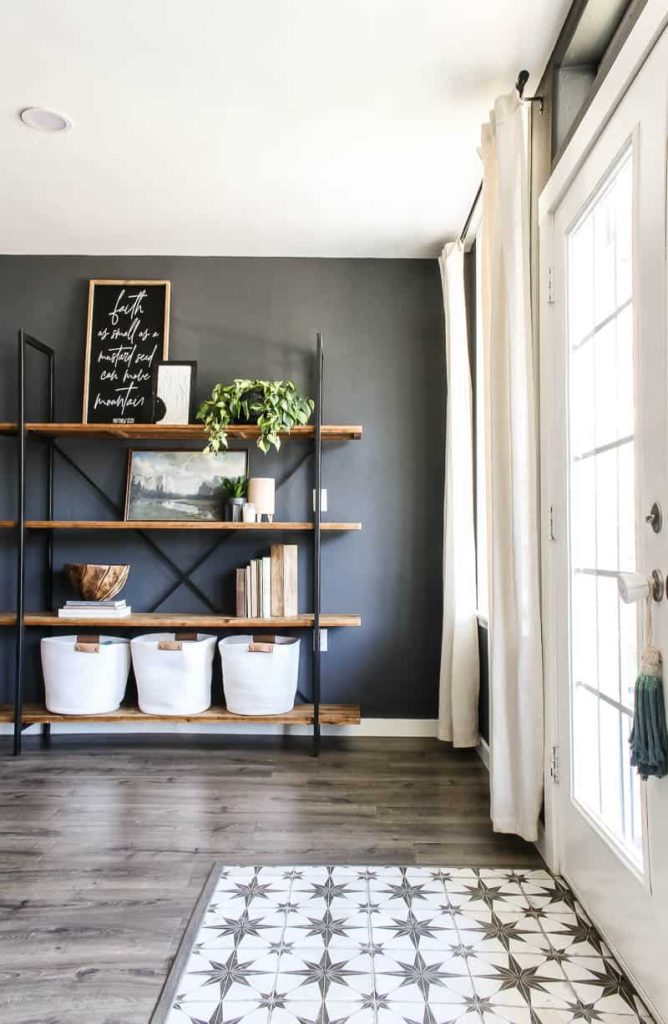 Image of metal bookshelf in modern boho living room with four wooden shelves minimally styled with books, greenery, white fabric baskets with leather handles, and artwork against dark grey wall.