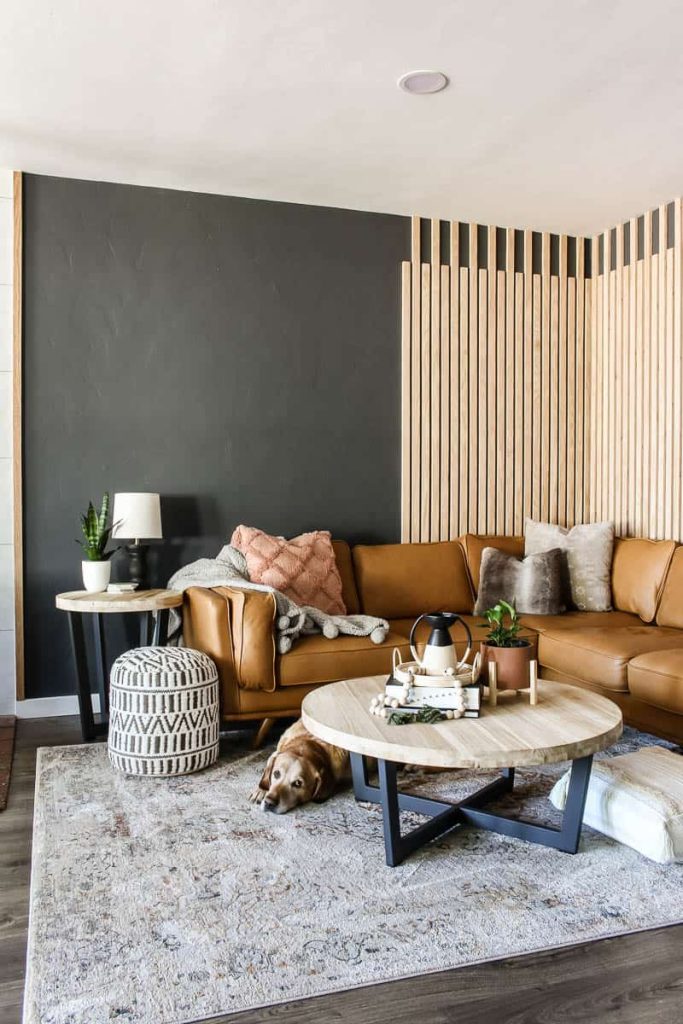 The before and afters of this modern boho living room renovation. Closeup of seating area with light brown cognac leather sectional, coffee table, wood slat accent wall and a lab looking up from between the sectional and coffee table styled with modern boho decor 