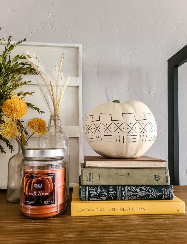 A photo of a white-painted pumpkin with mudcloth patterns drawn onto it using a Sharpie pen and a book for a cheap fall decoration.