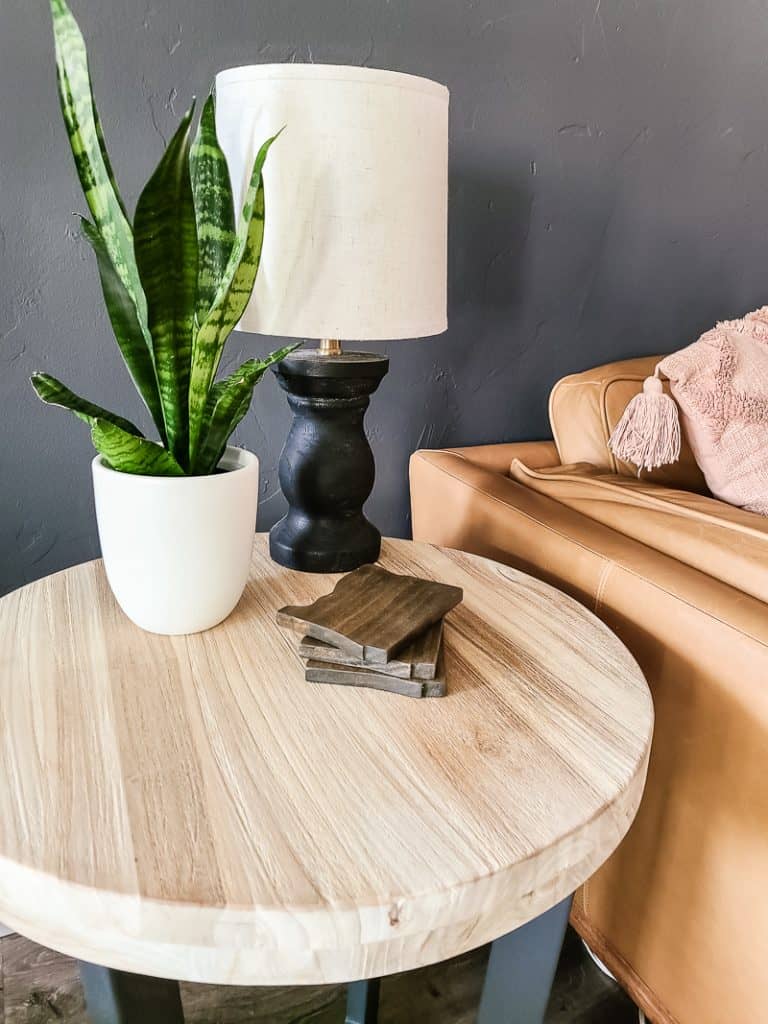 A pile of three dark-stained wooden coasters sit on a light wooden tabletop, next to a potted plant and lamp.