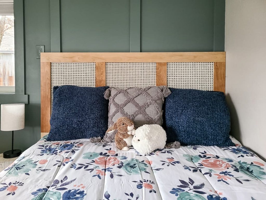 Close up front view of the bed with floral bed spread, blue and grey pillows, bunny and sheep stuffed toys and the diy cane headboard