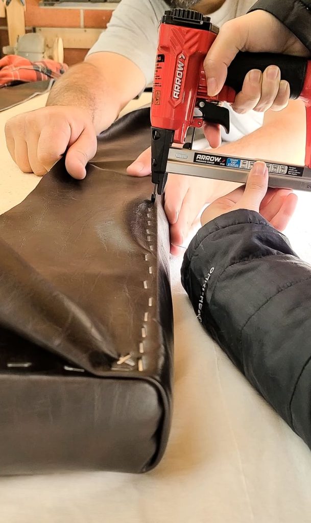 Man's hands pulling leather material tight so woman can staple leather fabric to the DIY bench cushion with an Arrow staple gun.