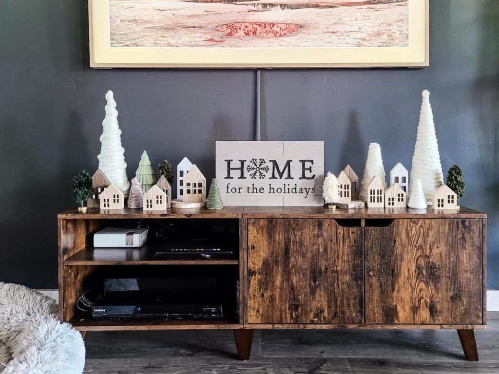 Mini Christmas village made up of wooden houses and diy yarn Christmas trees and a "Home for the holidays" wood sign at the center on top of the console table