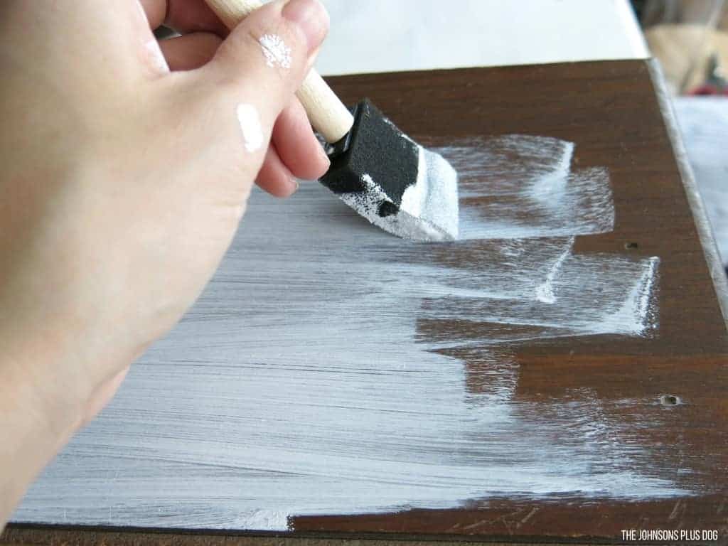 A person uses a small sponge brush to apply white chalk paint primer to a dark wooden surface.