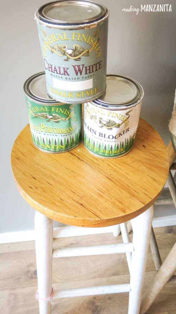 Three cans of chalk paint stacked on a wooden table. Each can has a different finish -- chalk style, satin, and white primer paint.