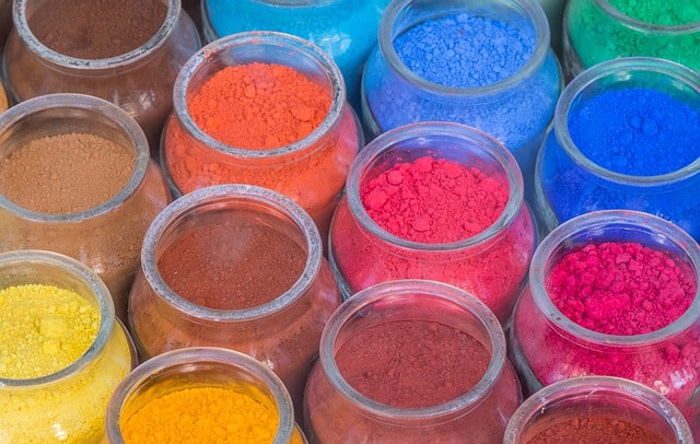 Glass jars are lined up in rows, filled with colorful powdered pigments in shades of blue, orange, pink, red, brown, and yellow.