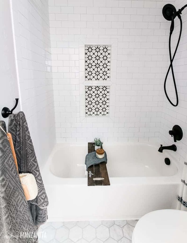 White tiled tub surround with black fixtures and bathtub tray with one shower niche within arms reach and the second directly above it at standing shower niche height.