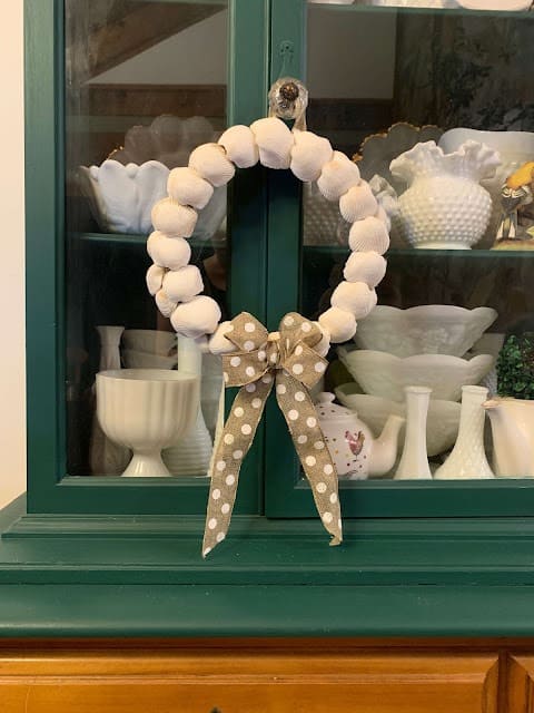 Wreath made using shells and tan polkadot ribbon hanging on doors of green cabinet
