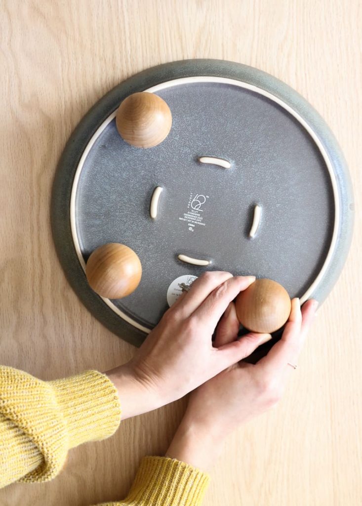 Overhead view of woman attaching third wood knob to bottom of ceramic plate to make the DIY footed tray.