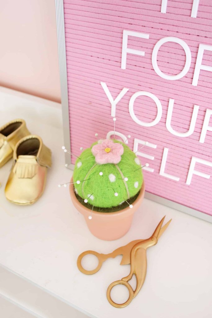 felted cactus shaped pin cushion with a flower on top next to gold scissors and gold baby shoes