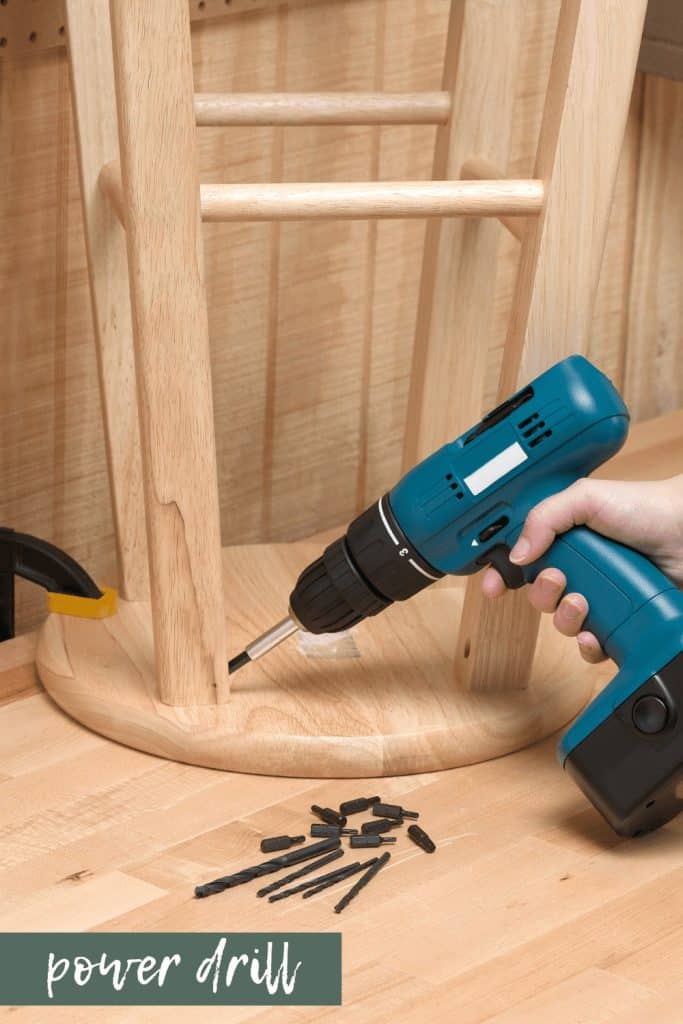 A person uses a blue power drill to insert metal screws into the leg of a wooden bar stool.