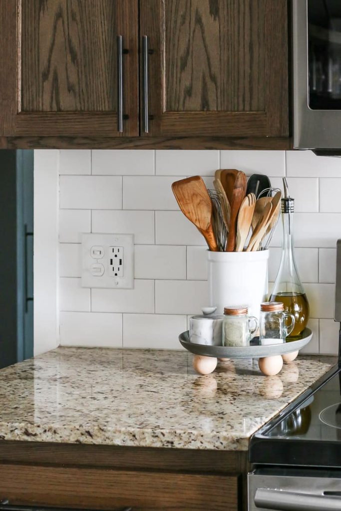 Close up of DIY footed tray on kitchen counter next to stove with kitchen utensils in white container, oil in dispenser, salt and pepper shakers and small container with lid.