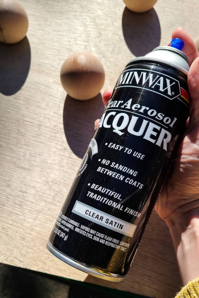 Woman's hand holding can of Minwax clear aerosol lacquer with three wood knobs laying on piece of wood.