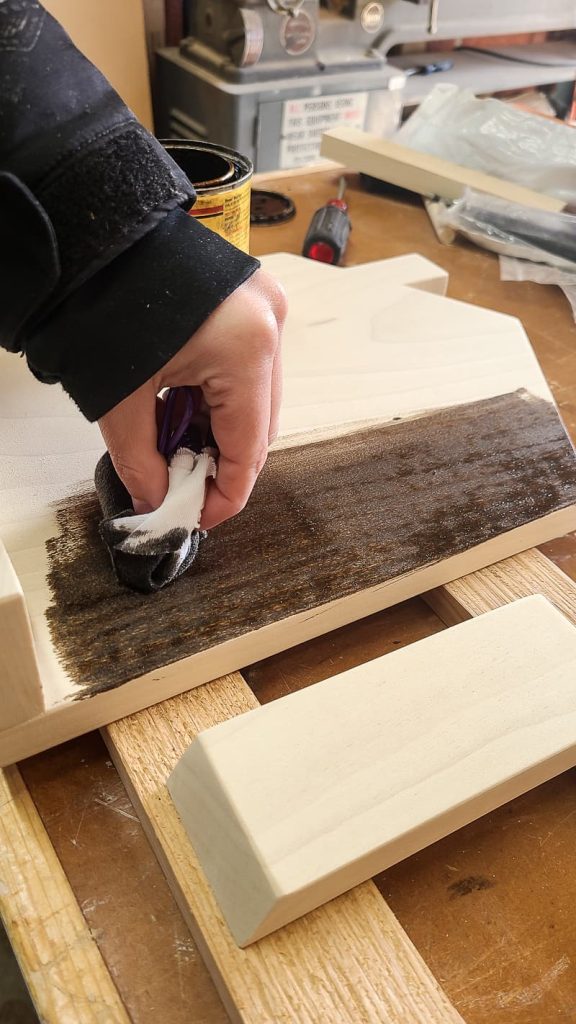 Image of woman's hand applying wood stain to a piece of wood using a rag.