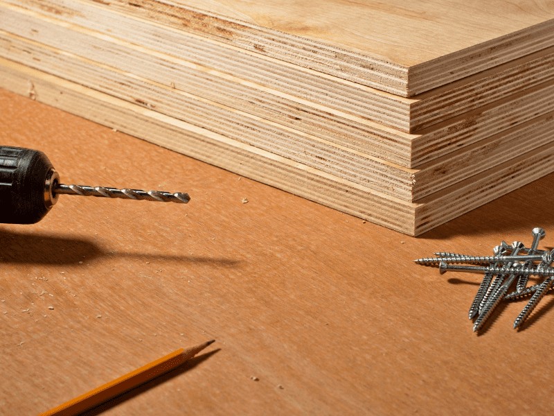 Image of a stack of plywood, screws, a pencil and the end of a drill with a drill bit.
