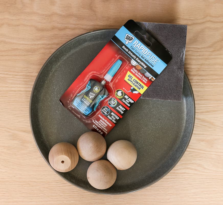 Supplies for DIY footed tray, ceramic plate, adhesive, sanding block, lacquer, and wooden knobs.