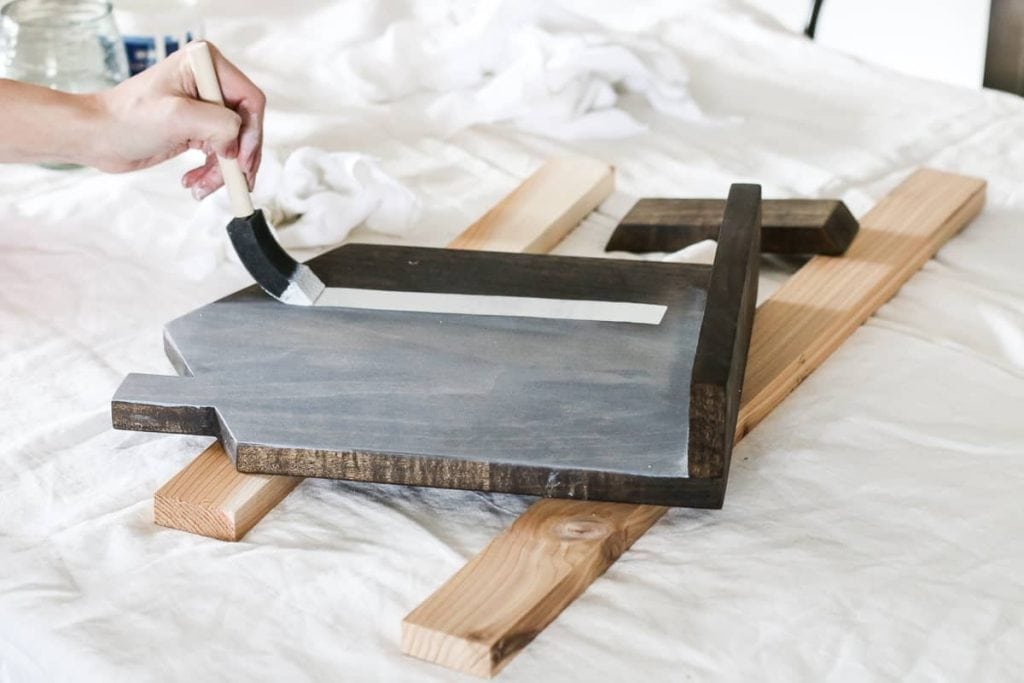 Woman's hand applying watered down white paint over stain with a sponge paintbrush on the wood cookbook stand sitting on two pieces of scrap wood on top of paint cloth.