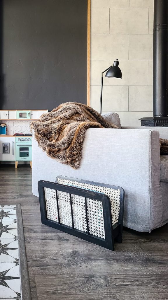 A thrifted magazine holder sits on a hardwood floor, next to an armchair. The wooden magazine holder is stained a dark black with light colored cane webbing on the inside.