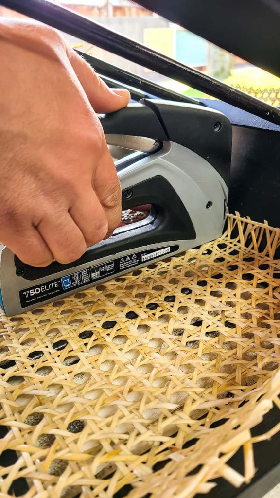 A person uses a staple gun to attach cane webbing to the inside of a wooden magazine rack.