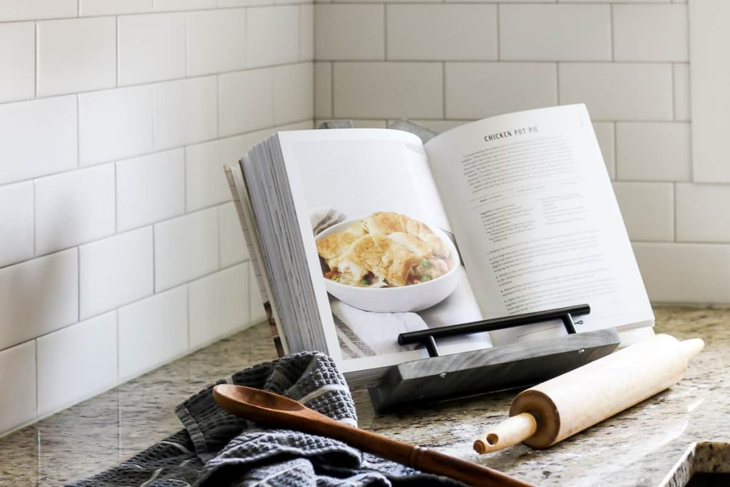 Cookbook open to recipe of chicken pot pie on the DIY cookbook stand with a rolling pin and wooden spoon and gray kitchen towel sitting on the kitchen counter.