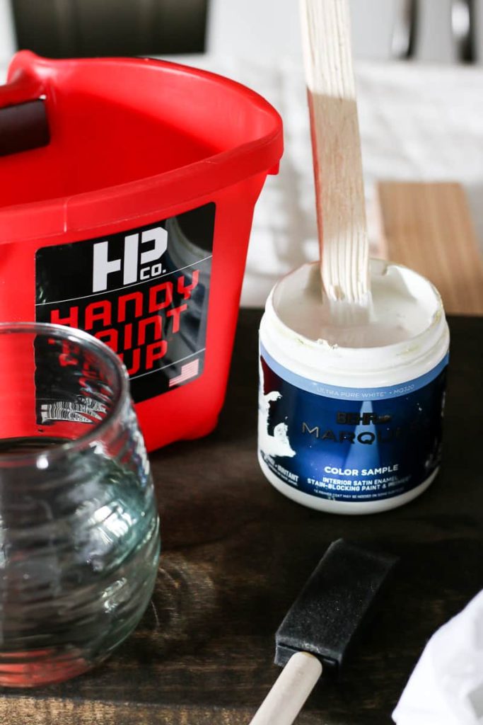 Small container of Behr Marquee in white with a paint stick in it, a Handy paint cup, clear glass container and sponge paintbrush sitting on a work surface.
