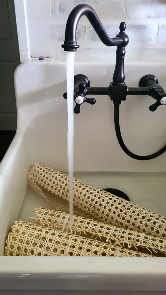 Rolls of cane webbing sit in a sink, filling with running water from a black faucet.