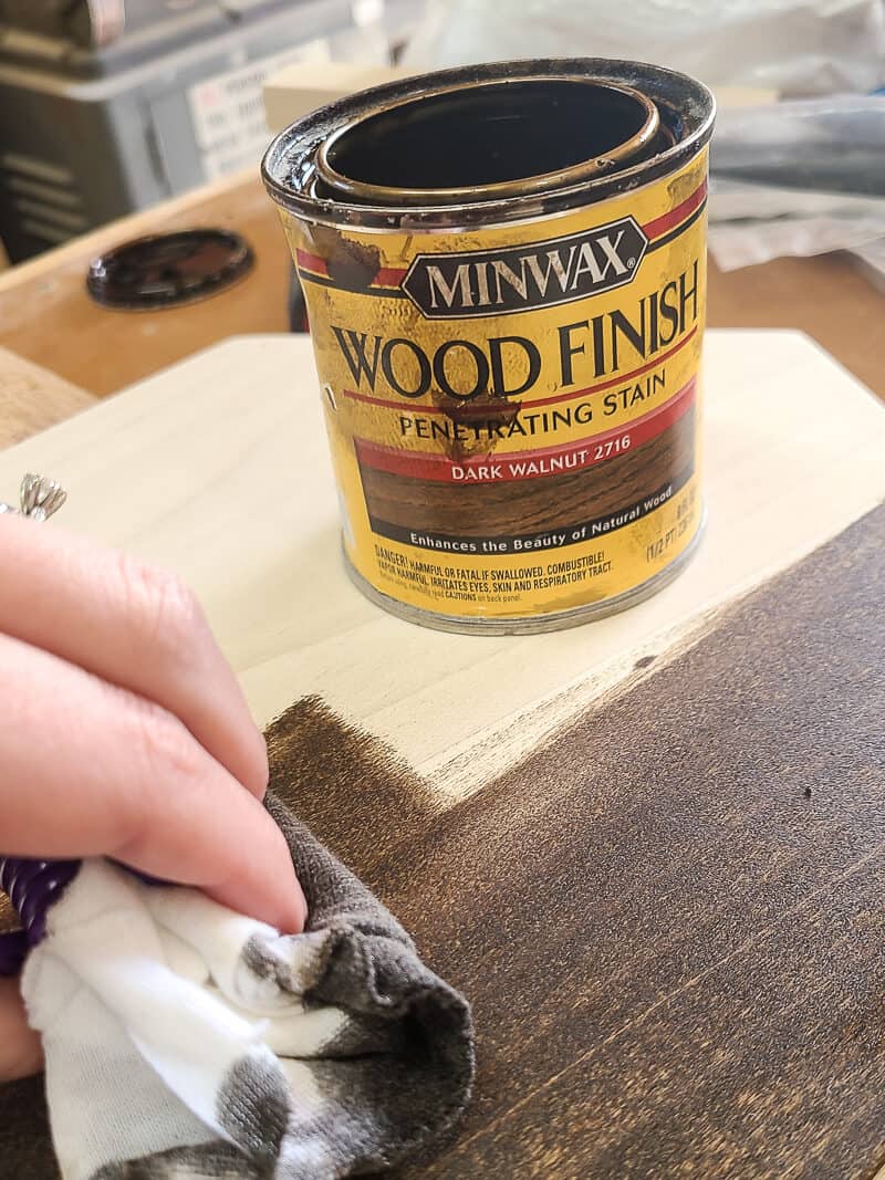 Can of Minwax wood finish in dark walnut sitting on top of piece of wood with a woman's hand applying the stain using a white rag.
