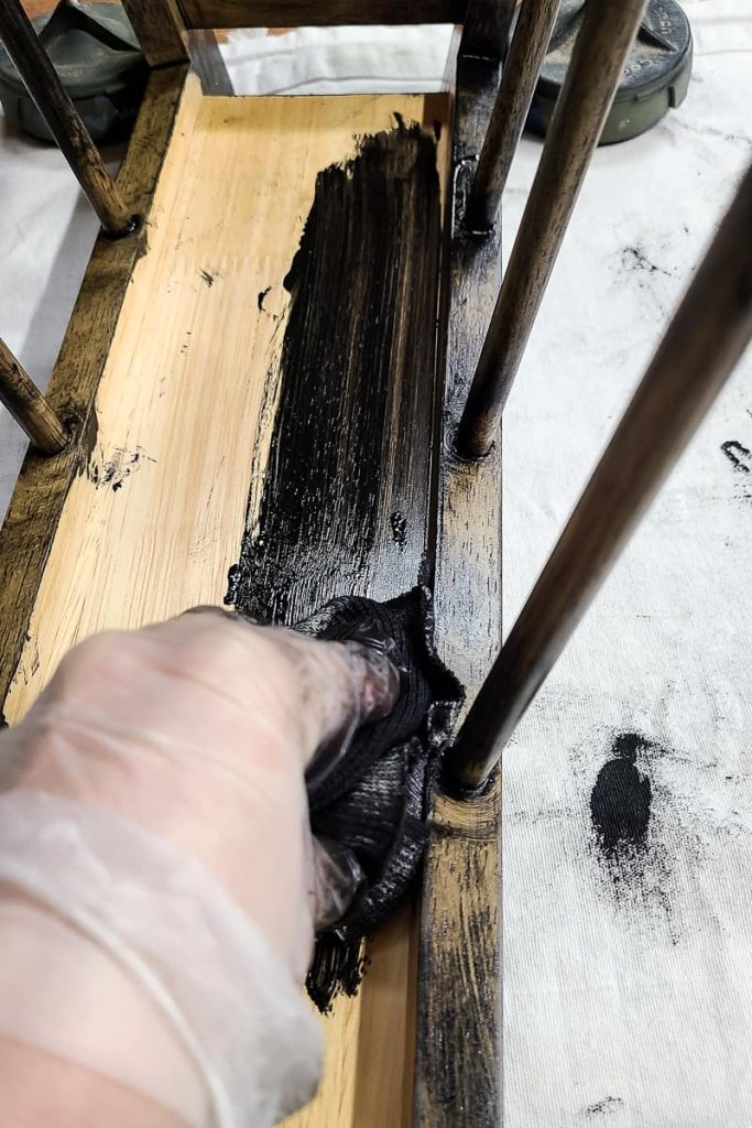 A person applies a black wood stain to an old thrifted magazine holder. The person is wearing gloves and using an old rag to apply the stain.