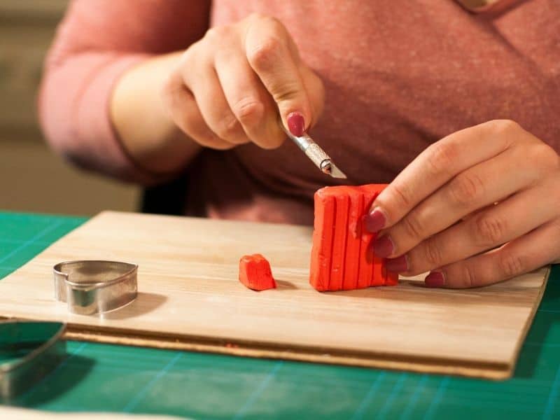 Woman cutting a brick of red polymer clay on a scrap piece of flooring with a heart shaped cookie cutter laying next to it.