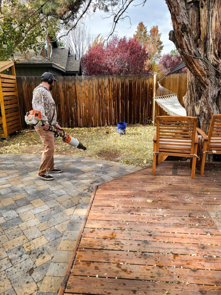 Man using a leaf blower to clean off leaves from deck and patio so they don't damage the deck or pavers during the winter season