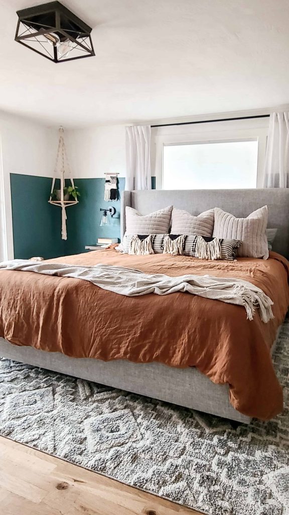 A king size bed dressed in brand new bohemian style bedding -- a rust colored linen duvet cover with a cream colored throw blanket at the foot of the bed, and two rows of accent pillows at the head of the bed.