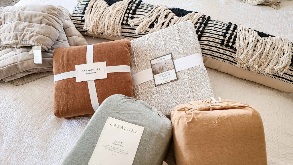 A collection of brand new bohemian styled bedding. Two sets of bed sheets, a rust colored duvet cover, cream throw blanket, and black and white patterned throw pillows.