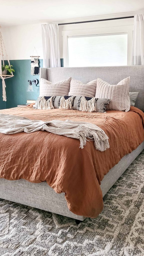 A large king-size bed is made up with neutral bohemian style bedding. The gray upholstered headboard supports rows of fluffy pillows, and the bed is covered with a rust colored duvet and cream throw blanket at the foot of the bed.