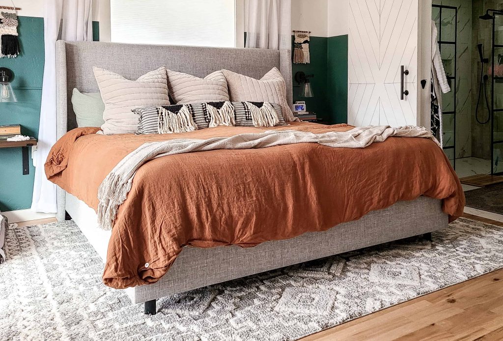 A king-size bed made up with a terracotta colored duvet cover, cream blanket, and rows of pillows against the upholstered headboard. The large bed sits on an area rug in a spacious master bedroom.