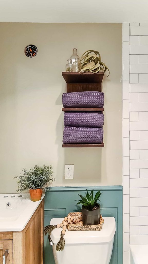 This dark wood stained wooden wall mounted towel shelf is the perfect place to store your clean and folded towels for guests over the toilet hanging on the wall. 